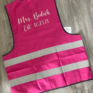 Op de afbeelding: Een felroze veiligheidsvest met twee reflecterende zilveren strepen. Het vest heeft een zwarte rand en de tekst "Mrs. Bedich Est. 10.23.21" in witte letters. Een klein label met tekst is zichtbaar aan de rechterkant.