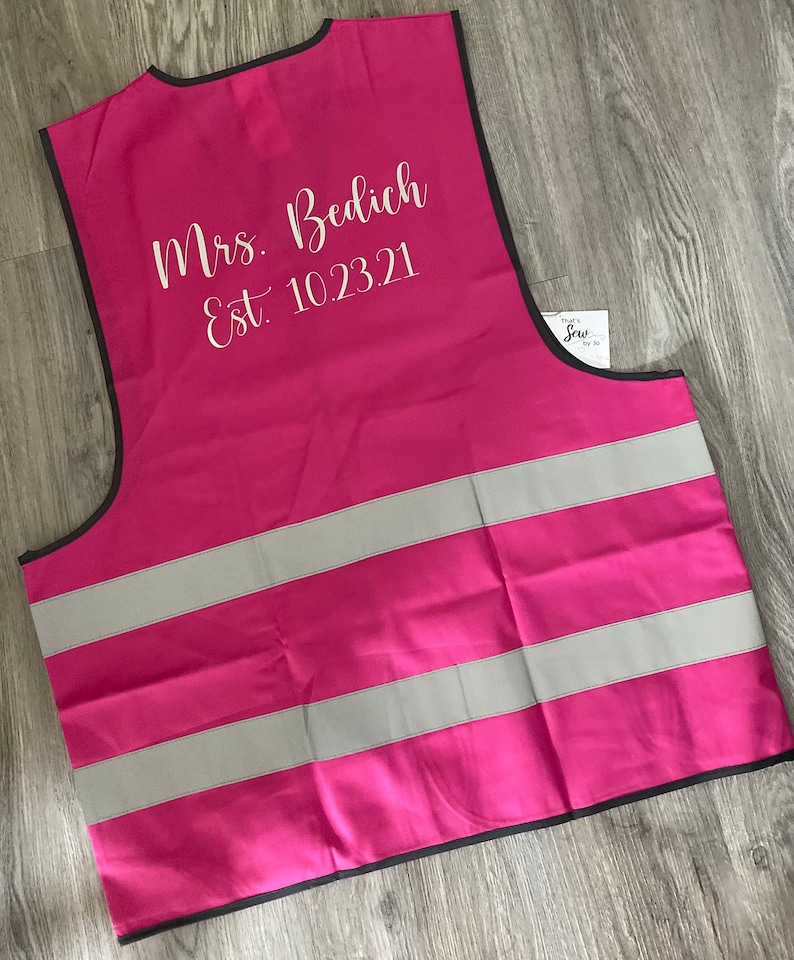 Op de afbeelding: Een felroze veiligheidsvest met twee reflecterende zilveren strepen. Het vest heeft een zwarte rand en de tekst "Mrs. Bedich Est. 10.23.21" in witte letters. Een klein label met tekst is zichtbaar aan de rechterkant.