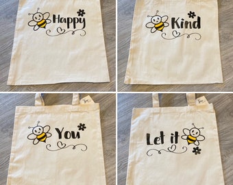May include: Four white tote bags with black bee and flower designs and text. The bags read "Happy", "Kind", "You", and "Let it".