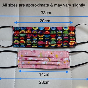 May include: Two face masks with black elastic ear loops. The top mask has a black background with colorful polka dots and measures 33 cm wide and 20 cm tall. The bottom mask has a pink background with a unicorn and rainbow pattern and measures 28 cm wide and 14 cm tall.
