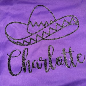 Op de afbeelding: Paars textiel met een glinsterend zwart sombrero-ontwerp en de naam "Charlotte" in een sierlijk lettertype. Het ontwerp is gecentreerd, met een witte rand aan de onderkant.