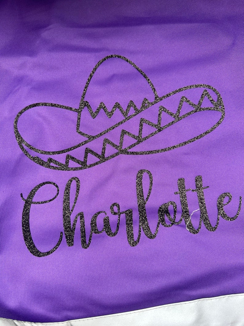 Op de afbeelding: Paars textiel met een glinsterend zwart sombrero-ontwerp en de naam "Charlotte" in een sierlijk lettertype. Het ontwerp is gecentreerd, met een witte rand aan de onderkant.