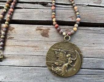 A round pendant with a leaf fairy on a crescent moon in an antiqued gold look hung from a strand of jasper beads.