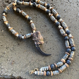 Peut inclure: Un collier composé de perles rayées noires et blanches, de perles en bois brun et de perles de lave noire. Le collier a un grand pendentif en forme de griffe brun foncé.