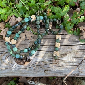 May include: A green and white beaded necklace with bone accents. The necklace is made of natural materials and has a rustic, earthy feel.