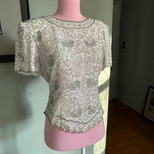 May include: A white, short-sleeved top with intricate silver beading and embellishments. The round neckline and hemline are also trimmed with silver beads. The top is displayed on a pink dress form, showcasing its delicate floral and leaf patterns.