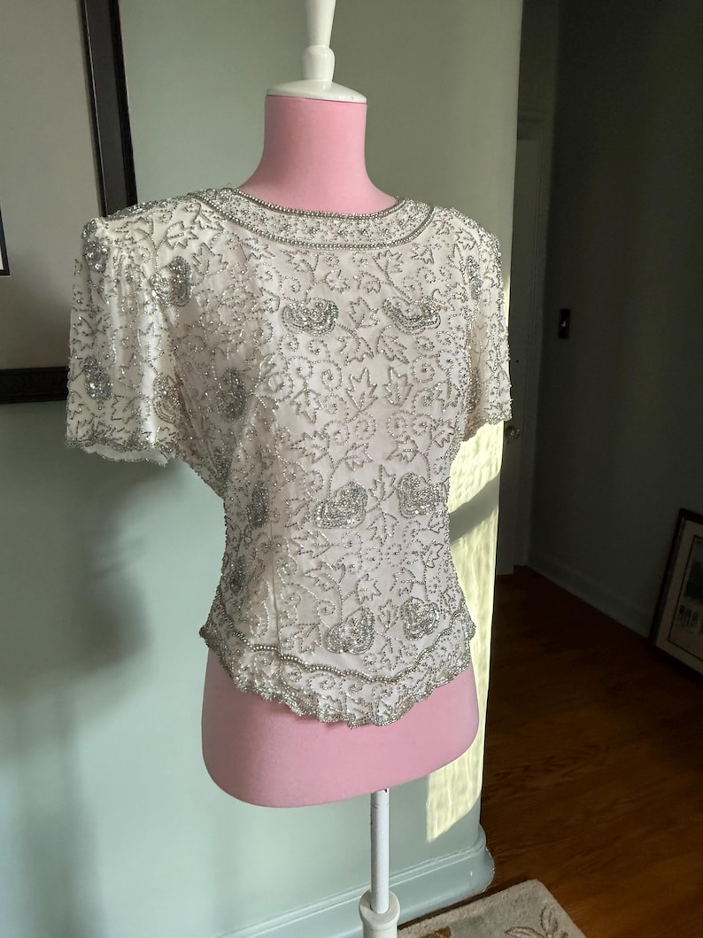 May include: A white, short-sleeved top with intricate silver beading and embellishments. The round neckline and hemline are also trimmed with silver beads. The top is displayed on a pink dress form, showcasing its delicate floral and leaf patterns.