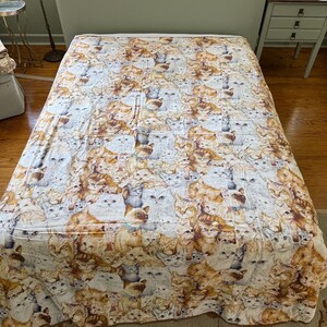 May include: A white bedspread featuring a pattern of numerous cat faces in various shades of brown, white, and grey.