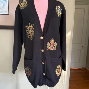 May include: Black cardigan sweater with gold buttons and embroidered crests. The long-sleeved sweater has a V-neck and two front pockets. The crests are in gold, red, and green.