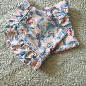 May include: A folded, floral-patterned bed sheet with a ruffled edge. The sheet features a light pink background with blue ribbons and floral designs in shades of pink, green, and blue. The ruffled edge is trimmed with a light blue piping.