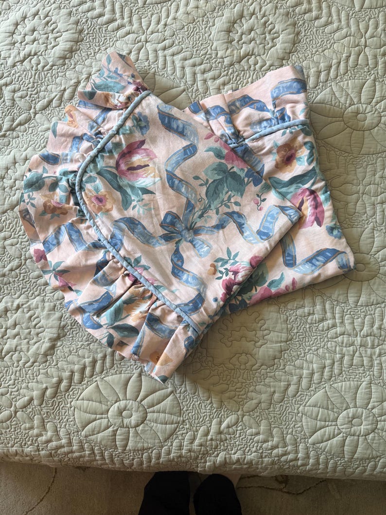 May include: A folded, floral-patterned bed sheet with a ruffled edge. The sheet features a light pink background with blue ribbons and floral designs in shades of pink, green, and blue. The ruffled edge is trimmed with a light blue piping.