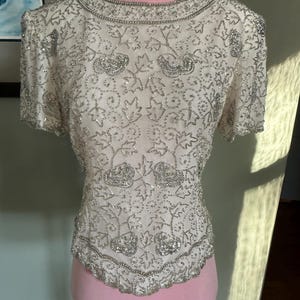 May include: Ivory-colored, short-sleeved top with intricate silver beadwork. The top features a round neckline and a scalloped hem, with floral and leaf patterns throughout. The garment is displayed on a pink mannequin.