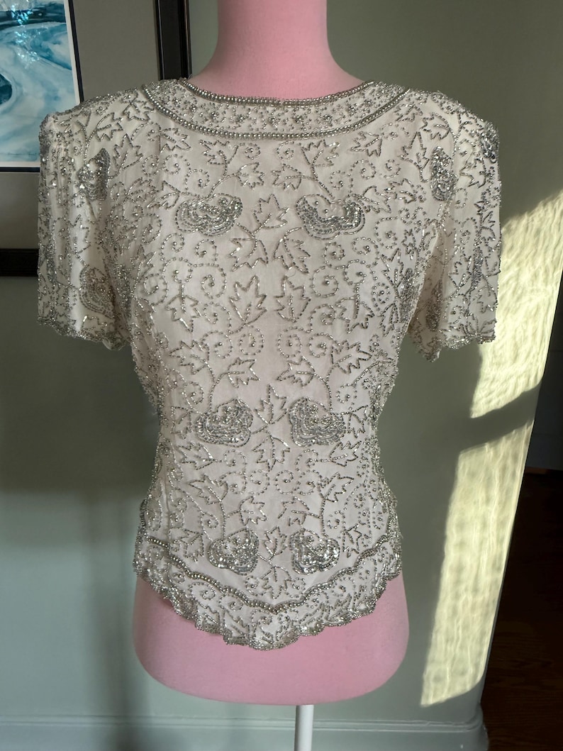 May include: Ivory-colored, short-sleeved top with intricate silver beadwork. The top features a round neckline and a scalloped hem, with floral and leaf patterns throughout. The garment is displayed on a pink mannequin.