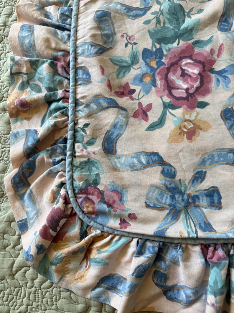 May include: Close-up of a floral pillowcase with a ruffled edge. The fabric features a pattern of pink, blue, and green flowers and ribbons on a cream background. A blue cord trim frames the ruffled edge.