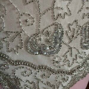 May include: Close-up of a cream-colored fabric adorned with intricate silver beadwork. The design features floral and abstract patterns, with the beads creating a shimmering, textured surface. The detailed embroidery suggests a vintage or handmade item.