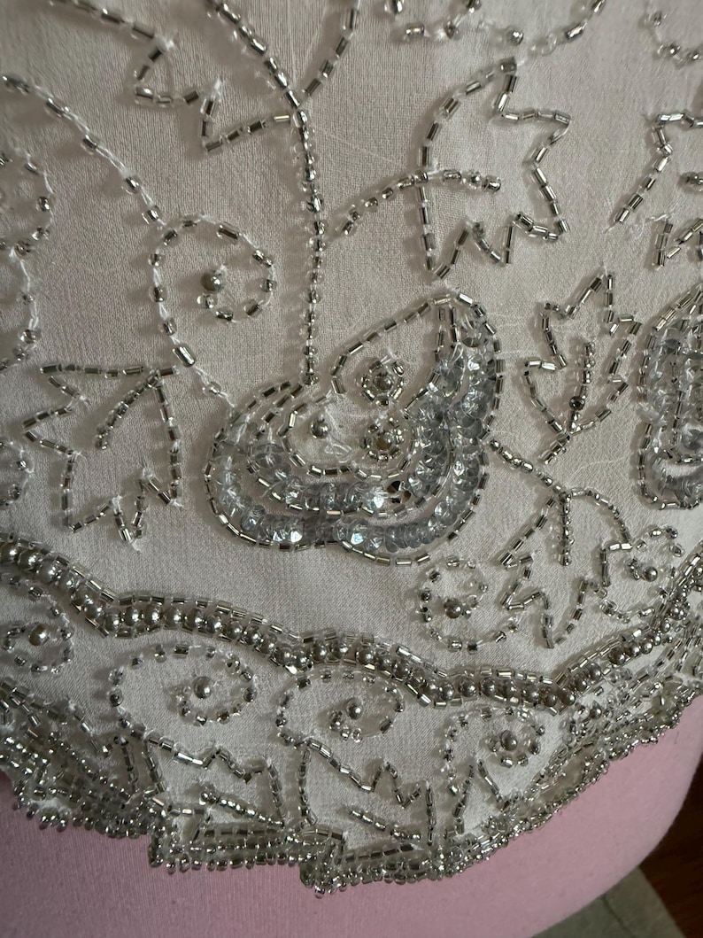 May include: Close-up of a cream-colored fabric adorned with intricate silver beadwork. The design features floral and abstract patterns, with the beads creating a shimmering, textured surface. The detailed embroidery suggests a vintage or handmade item.