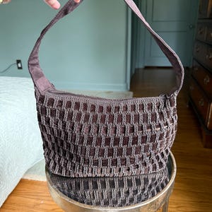 May include: A brown shoulder bag with a woven design. The bag has a rectangular shape with a zipper closure and a matching shoulder strap. The bag is made of a soft, textured material.