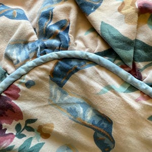 May include: Close-up of a floral patterned fabric in shades of pink, blue, green, and yellow. The design features large blue ribbons and floral elements. A light blue cord trims the edge of the fabric.