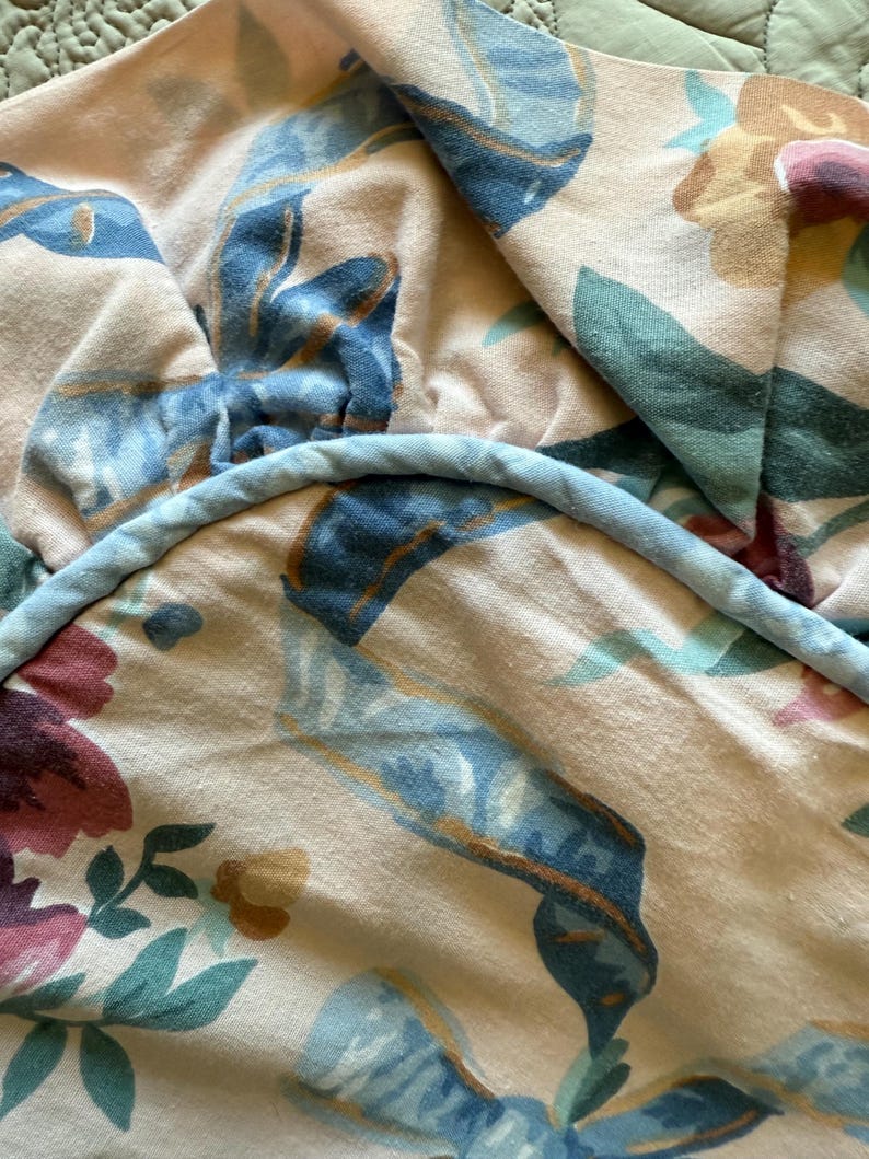May include: Close-up of a floral patterned fabric in shades of pink, blue, green, and yellow. The design features large blue ribbons and floral elements. A light blue cord trims the edge of the fabric.