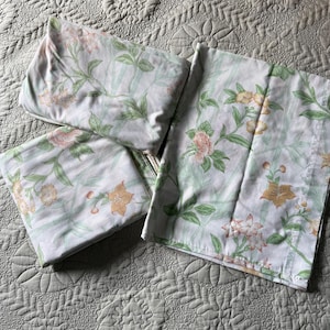 Puede incluir: Un juego de sábanas blancas con un estampado floral de flores rosas y amarillas y hojas verdes. Las sábanas están dobladas y colocadas sobre un cubrecama acolchado.