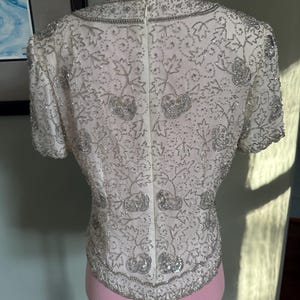 May include: Ivory-colored, short-sleeved top with intricate silver beading and sequin embellishments. The design features floral and leaf patterns, with a beaded collar and hemline. The top is displayed on a pink mannequin.