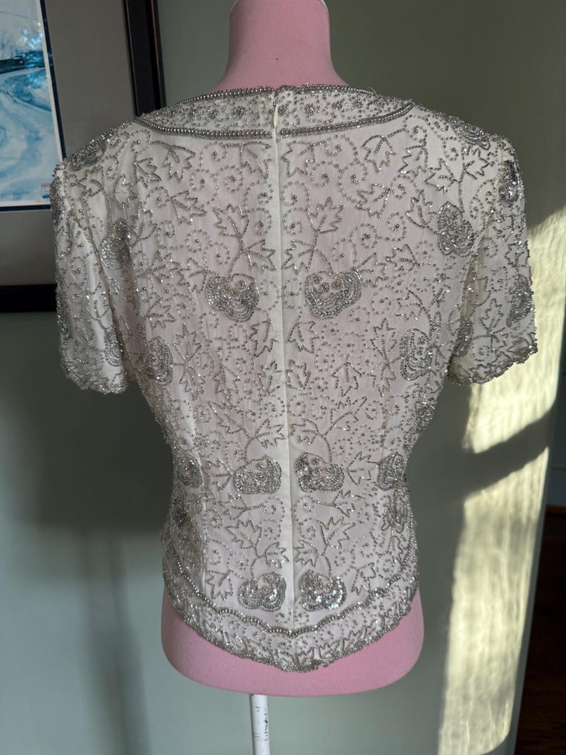 May include: Ivory-colored, short-sleeved top with intricate silver beading and sequin embellishments. The design features floral and leaf patterns, with a beaded collar and hemline. The top is displayed on a pink mannequin.