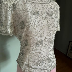 May include: Ivory-colored, short-sleeved top with intricate silver beadwork. The design features floral and leaf patterns, with a beaded neckline and hem. The top is displayed on a pink mannequin, showcasing its detailed embellishments.