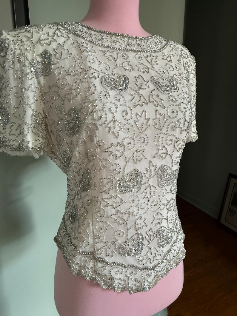 May include: Ivory-colored, short-sleeved top with intricate silver beadwork. The design features floral and leaf patterns, with a beaded neckline and hem. The top is displayed on a pink mannequin, showcasing its detailed embellishments.
