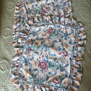 May include: Two decorative pillow shams with a floral and ribbon pattern. The shams feature a light pink background with blue ribbons and colorful flowers. Each sham has a ruffled edge and a blue trim.
