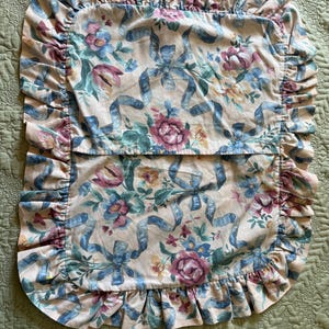 May include: A floral pillowcase with a ruffled edge. The pillowcase features a pattern of blue ribbons, pink roses, and other flowers on a cream background. The ruffled edge is a light pink color.