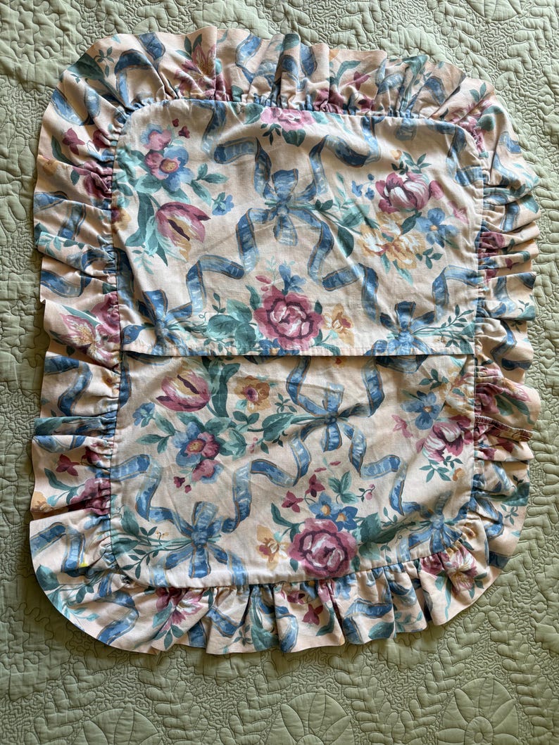 May include: A floral pillowcase with a ruffled edge. The pillowcase features a pattern of blue ribbons, pink roses, and other flowers on a cream background. The ruffled edge is a light pink color.