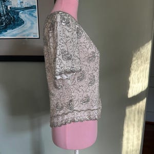 May include: White, short-sleeved top with silver sequin and bead embellishments. The top is displayed on a pink dress form. The design features floral patterns and a beaded hemline, creating a shimmering effect. The sleeves have a white trim.