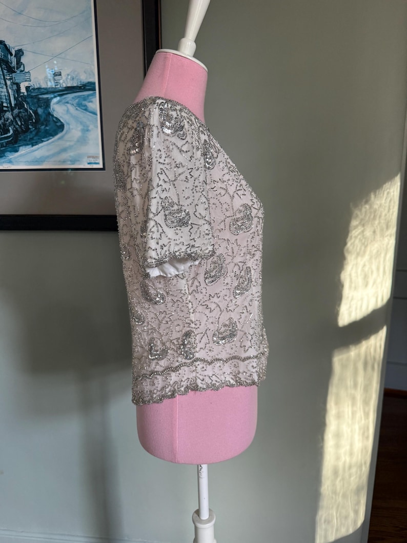 May include: White, short-sleeved top with silver sequin and bead embellishments. The top is displayed on a pink dress form. The design features floral patterns and a beaded hemline, creating a shimmering effect. The sleeves have a white trim.