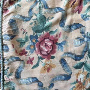 May include: Floral fabric featuring a repeating pattern of roses, tulips, and other flowers in shades of pink, blue, and yellow. Blue ribbons are interspersed throughout the design, set against a light peach background.