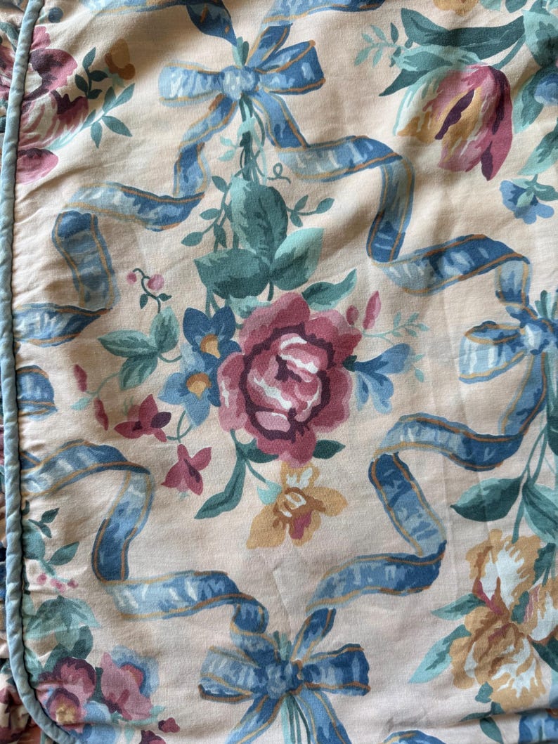 May include: Floral fabric featuring a repeating pattern of roses, tulips, and other flowers in shades of pink, blue, and yellow. Blue ribbons are interspersed throughout the design, set against a light peach background.