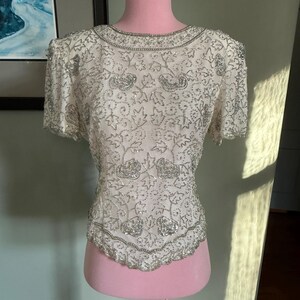 May include: Ivory-colored, short-sleeved top with intricate silver beading. The top features a round neckline and a scalloped hemline, with floral and leaf patterns. The garment is displayed on a pink mannequin, showcasing its design and texture.