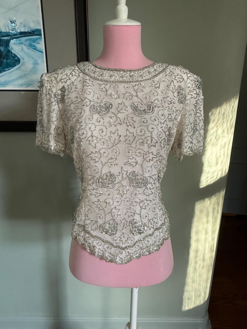 May include: Ivory-colored, short-sleeved top with intricate silver beading. The top features a round neckline and a scalloped hemline, with floral and leaf patterns. The garment is displayed on a pink mannequin, showcasing its design and texture.