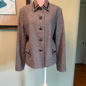 May include: A grey tweed blazer with black trim on the collar, pockets, and cuffs. The blazer has a classic design with a notched collar and four black buttons down the front. The jacket is displayed on a white mannequin.