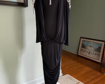 May include: A black dress with a draped bodice and a ruched skirt. The dress features embellished shoulder details and a decorative brooch. The dress is displayed on a white dress form.