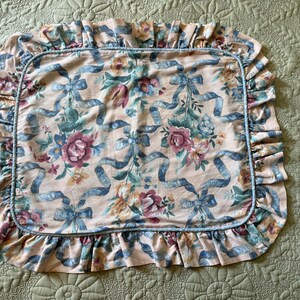 May include: A rectangular pillowcase with a floral and ribbon pattern. The pillowcase features a peach background with blue ribbons and various flowers in shades of pink, green, and yellow. The edges are trimmed with a ruffled border.