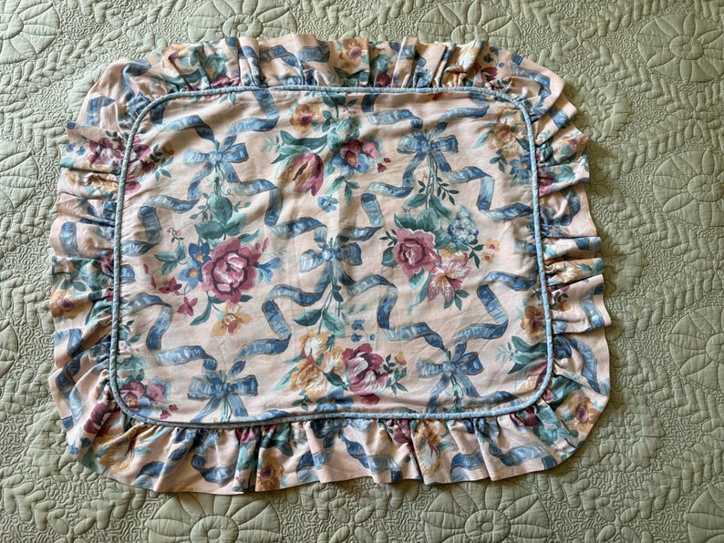 May include: A rectangular pillowcase with a floral and ribbon pattern. The pillowcase features a peach background with blue ribbons and various flowers in shades of pink, green, and yellow. The edges are trimmed with a ruffled border.