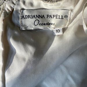 May include: Close-up of a white dress label with the text "ADRIANNA PAPELL Occasions" and the number "10". The dress appears to be made of a soft, flowing fabric, with beaded embellishments visible at the top.
