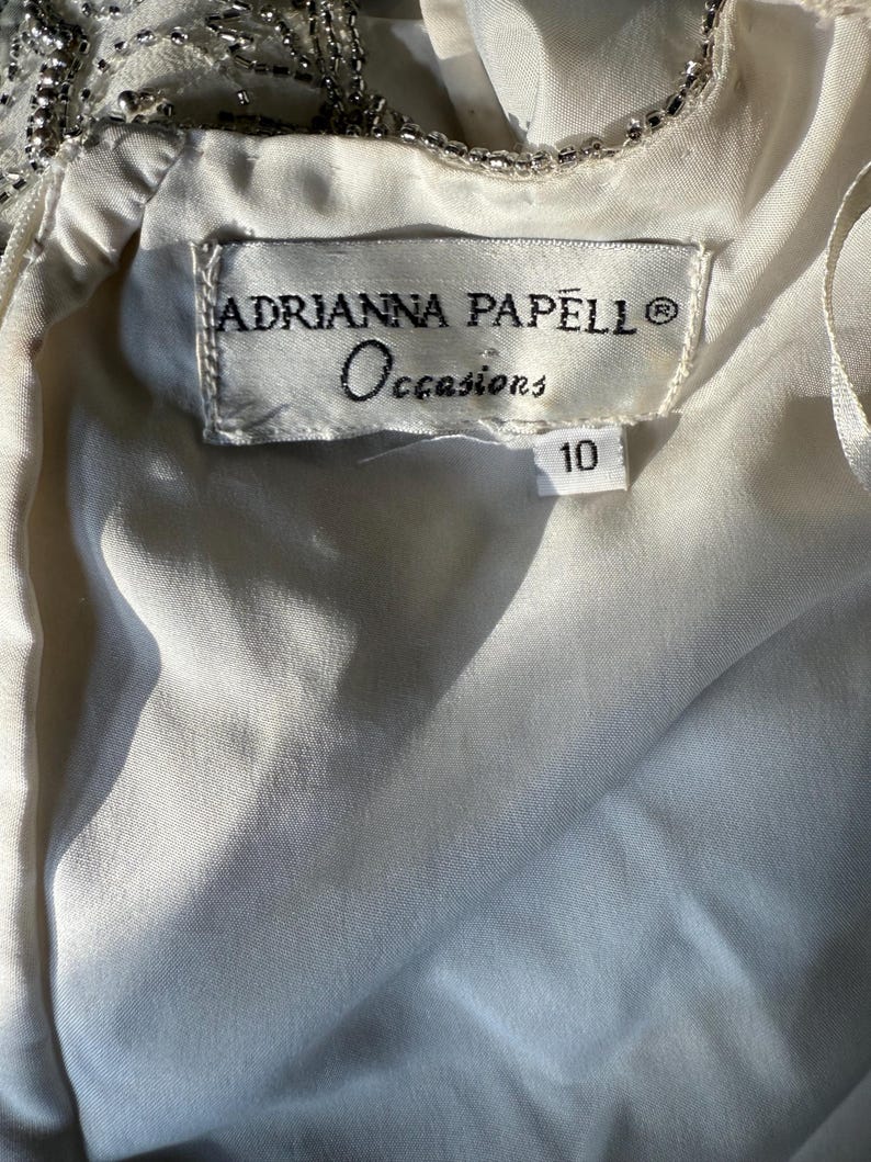 May include: Close-up of a white dress label with the text "ADRIANNA PAPELL Occasions" and the number "10". The dress appears to be made of a soft, flowing fabric, with beaded embellishments visible at the top.