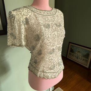 May include: Ivory-colored, short-sleeved top with intricate silver beading. The round neckline and hem are trimmed with silver beads. The bodice features floral and leaf patterns, creating a shimmering effect. The top is displayed on a pink mannequin.