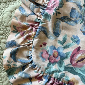 May include: Close-up of a floral patterned pillowcase with a ruffled edge. The design features pink, blue, and purple flowers with blue ribbons on a cream background. The pillowcase is on a green quilted surface.