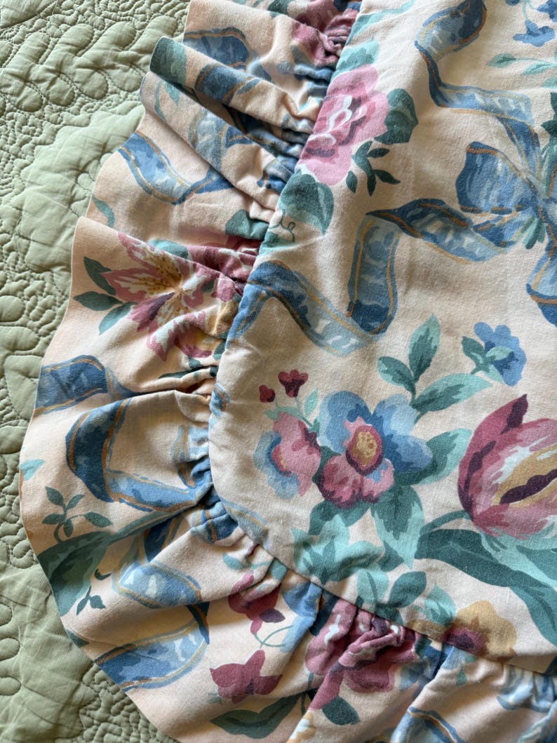 May include: Close-up of a floral patterned pillowcase with a ruffled edge. The design features pink, blue, and purple flowers with blue ribbons on a cream background. The pillowcase is on a green quilted surface.