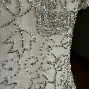 May include: Close-up of a white garment with intricate silver bead and sequin embellishments. The design features floral and abstract patterns, with the beads and sequins sewn onto the fabric in a detailed arrangement. The garment appears to be a dress or top.