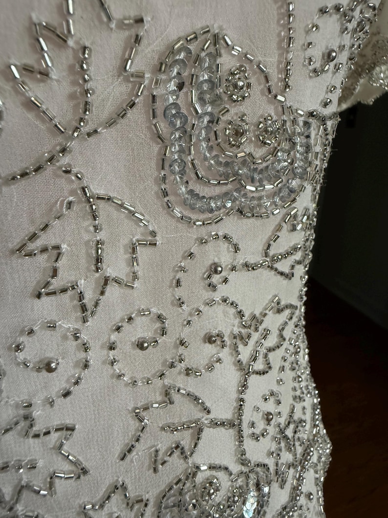 May include: Close-up of a white garment with intricate silver bead and sequin embellishments. The design features floral and abstract patterns, with the beads and sequins sewn onto the fabric in a detailed arrangement. The garment appears to be a dress or top.