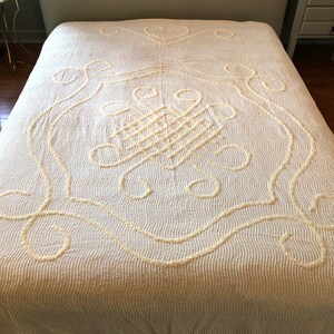 May include: A white crocheted bedspread with a large, intricate floral design. The bedspread has a fringe border and is made of a soft, fluffy material.