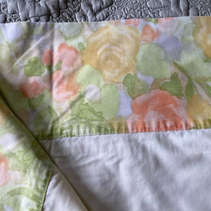 Może przedstawiać: Biała pościel z kwiatowym wzorem w odcieniach żółtego, zielonego i brzoskwiniowego. Wzór kwiatowy jest w stylu akwarelowym, z miękkim, wyblakłym wyglądem.